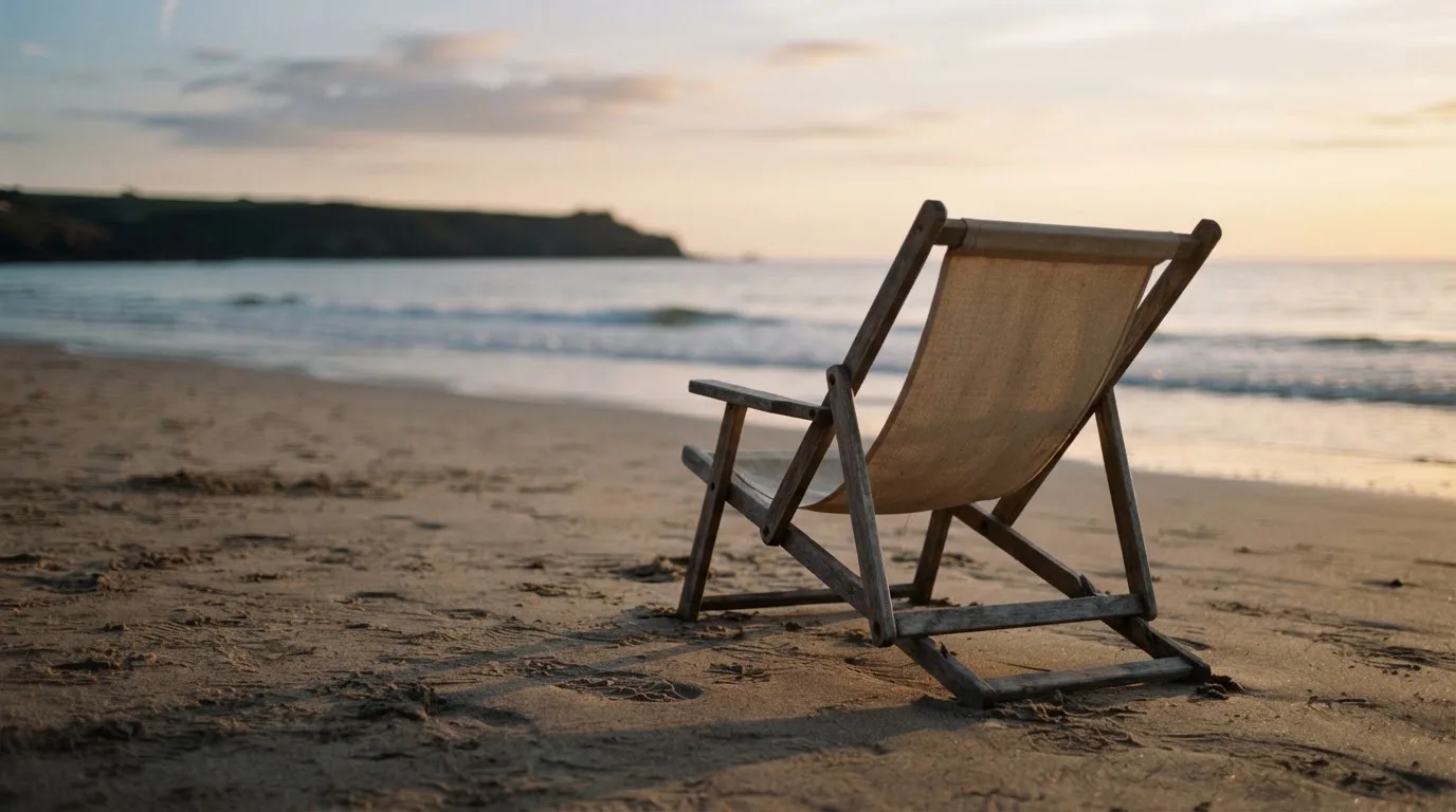 Ein leerer Liegestuhl aus Holz steht am Rand eines Sandstrands, Abendlicht und ruhige Wellen im Hintergrund.