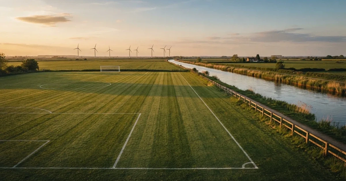 Ein Fussballplatz neben einem Kanal in flachem Land mit Windrädern am Horizont, Abendlicht liegt über dem Rasen