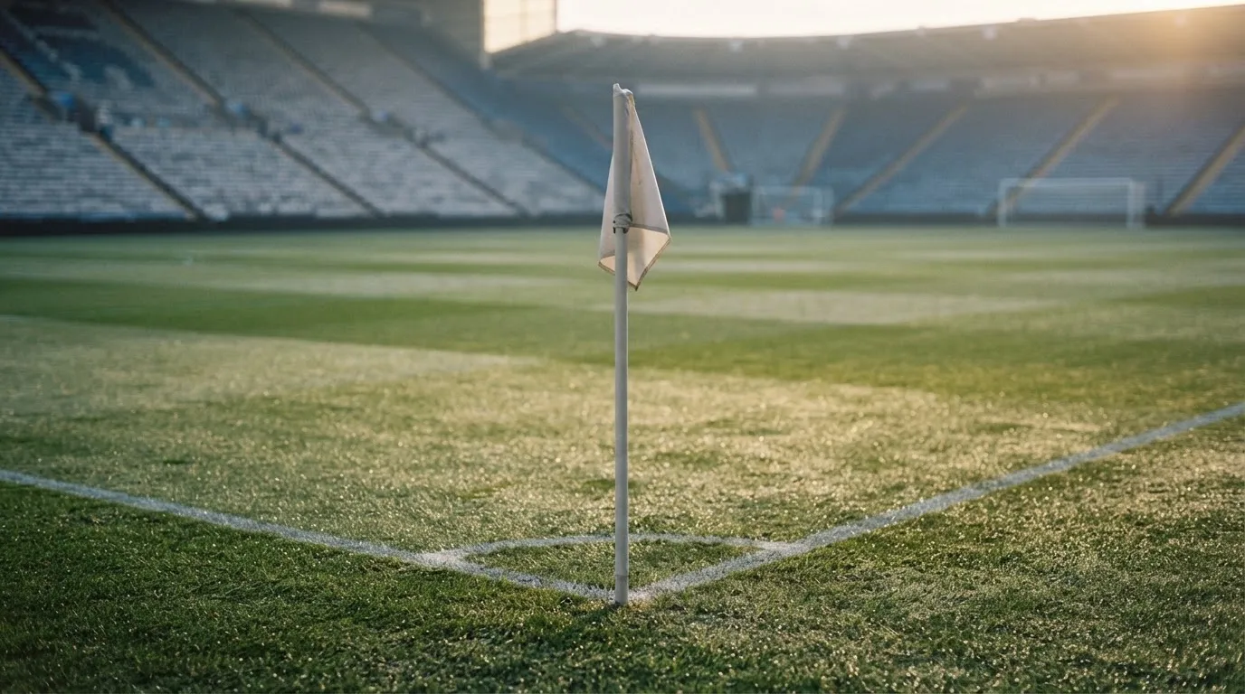 Eine Eckfahne steht auf nassem Rasen im ersten Morgenlicht, dahinter beginnt eine leere Tribüne.